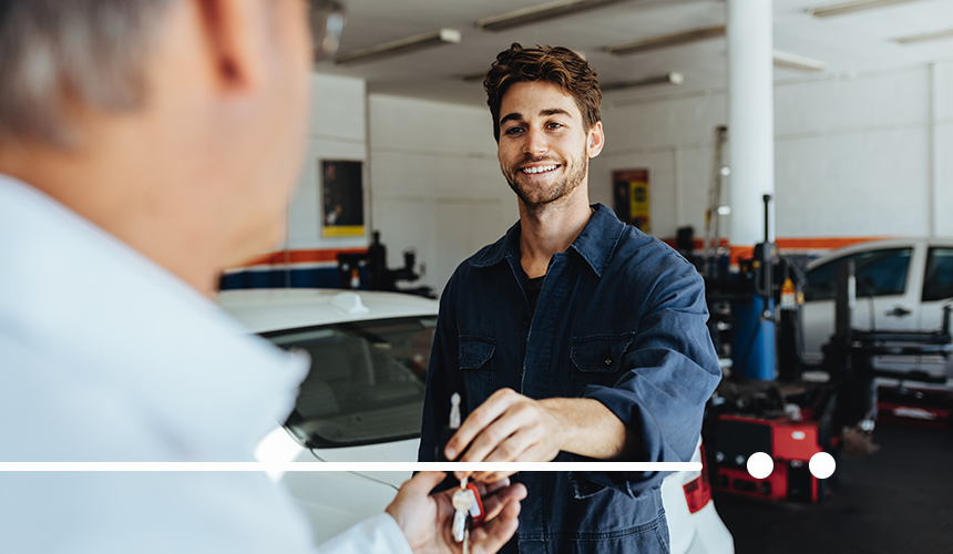 photo of workshop technician exchanging car keys with customer, white car visible in background, general setting of workshop