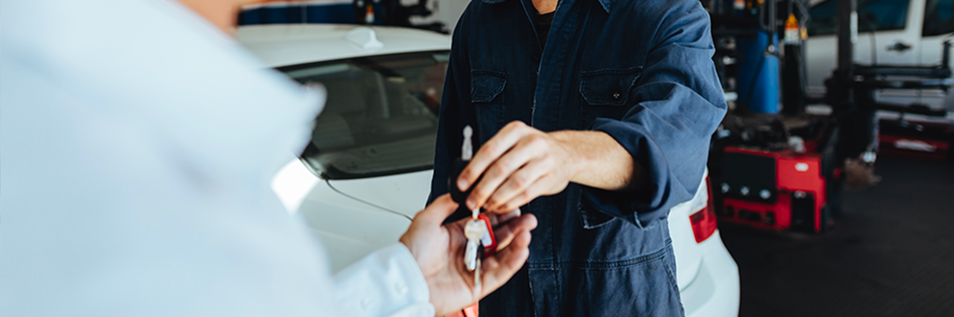 photo of workshop technician exchanging car keys with customer, white car visible in background, general setting of workshop
