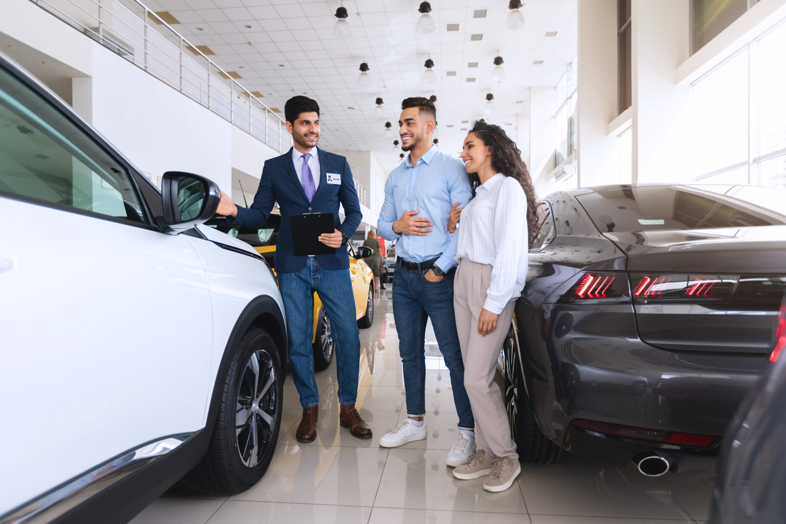 Three people stood in a car showroom between two cars. One person is a salesman, the other two are customers.