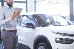 Salesman stood in a car dealership with one car behind him, he is typing on an ipad tablet style device