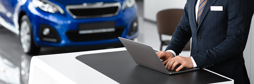 Person standing typing on a laptop on a desk, inside a car dealership, with the front of a car visible behind the desk